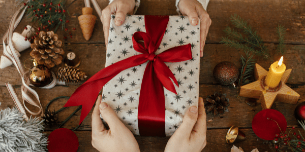 A giftbox wrapped in white paper with silver stars and a red box is being passed from one set of hands to another. On the wooden table under the gift there is a collection of festive items including pinecones, candles, bells, ribbon, and greenery.